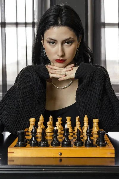 黒の美しいエレガントな少女の肖像画は 指が交差し 顎がそれらの上に残り 彼女の顔に深刻な表情でチェス盤の後ろに座って 熱心に見つめて — ストック写真