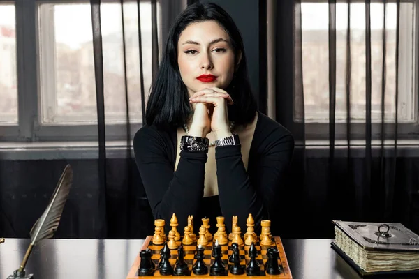 stock image Portrait of a beautiful, elegant girl in black, sits behind a chessboard with fingers crossed and chin resting on them and slightly smile expression on her face, staring intently.