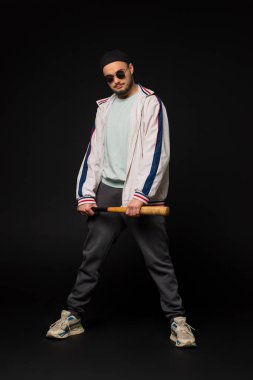 Sokakta eşofman ve yuvarlak güneş gözlüğü takmış elinde beyzbol sopasıyla gezen siyah bir arka planda izole edilmiş genç, sert bir adam. Sokak kültürü, sokak çetesi, şehir modası..