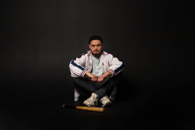 Sokakta eşofman giymiş genç, sert bir adam siyah bir arka planda beysbol sopasıyla yerde oturuyor. Şehir sokak kültürü.