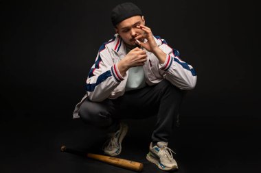 Sokakta eşofman giymiş çömelmiş sigara yakan genç, sert bir adam. Şehir sokak kültürü, sokak çetesi.