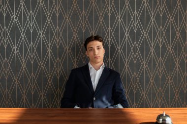 Resmi koyu mavi takım elbiseli genç bir adam servis masasının arkasında servis zili ile oturuyor. Hizmet personeli, profesyonel giyinmiş..