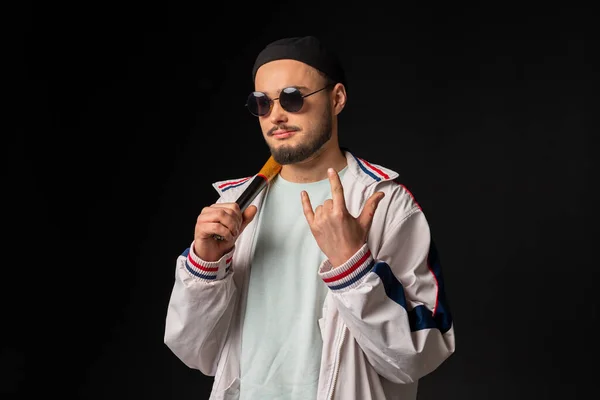 A young, tough guy from the streets dressed in a tracksuit and round sunglasses holding a baseball bat, posing, isolated on a black background. Urban street culture, Street gang, urban fashion.