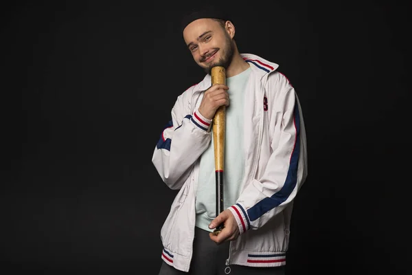 A young, tough but smiling guy from the streets dressed in a tracksuit holding a baseball bat, posing, isolated on a black background. Urban street culture, Street gang, urban fashion.
