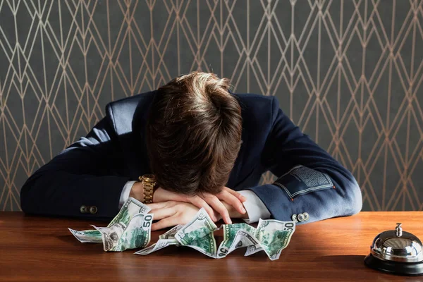 stock image The young man in a formal dark blue suit, looking disappointed and hopeless, rested his head on the table surrounded by crumpled money. A failed business, financial troubles, and a failed project.