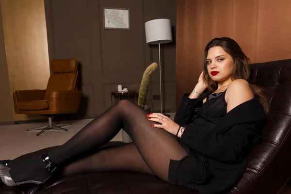 A young and attractive female secretary in stockings, formally and sensual dressed , sits in an office chair with loose hair, looking at the camera. Office decor and a tall cactus on a background.