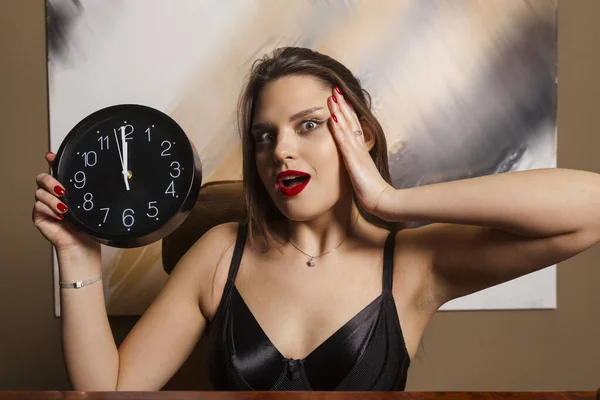 A young and attractive, surprized woman in an elegant black dress with long loose hair holding a watch. Concept of punctuality and time management.