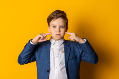 Mavi takım elbiseli ve beyaz gömlekli küçük bir çocuk, düzgün giyinmiş, ağzı sulanmış, parmaklarını yanaklarını işaret ediyor. Sarı arka planda izole edilmiş..