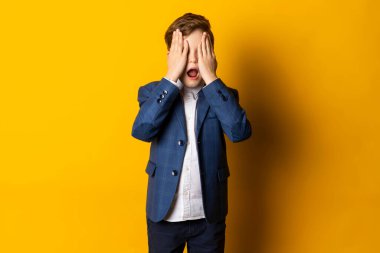 Mavi takım elbiseli ve beyaz gömlekli küçük bir çocuk, düzgün giyimli, elleriyle gözlerini kapatıyor ve bağırıyor..