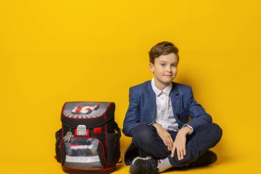 Mavi takım elbiseli, beyaz gömlekli, okul çantalı, düzgün giyimli, okul çantasının yanında oturan küçük bir çocuk. Sarıda izole edilmiş..
