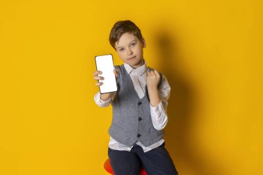 Gri yelek ve beyaz gömlekli mutlu bir çocuk. Temiz giyimli. Fotokopi odası için beyaz bir telefon ekranı gösteriyor. Sarı arkaplanda izole edilmiş.
