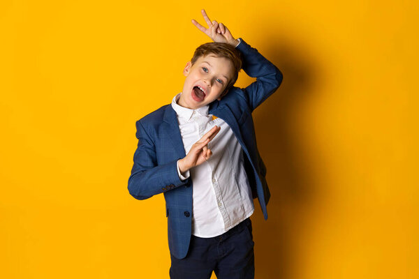 Счастливый маленький мальчик в синем костюме и белой рубашке, аккуратно одетый, полный радости, подражающий и жестикулирующий. Изолированный на желтом фоне.