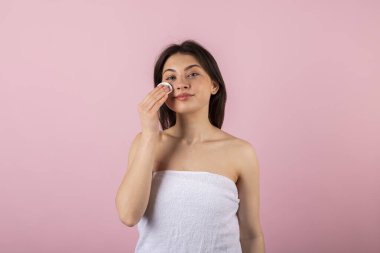 Sadece beyaz bir havluya sarılı çekici bir genç kız mutlu bir şekilde gülümsüyor ve pamuk veya ped kullanıyor. Pembe bir arkaplanda izole.