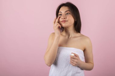 Sadece beyaz bir havluya sarılı çekici bir genç kız mutlu bir şekilde gülümsüyor ve yüzünde yeni bir yüz kremi kullanıyor. Pembe bir arkaplanda izole.