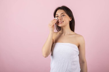 Sadece beyaz bir havluya sarılı çekici bir genç kız mutlu bir şekilde gülümsüyor ve pamuk veya ped kullanıyor. Pembe bir arkaplanda izole.