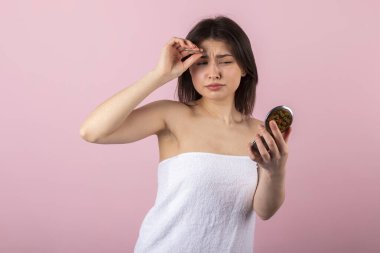 Sadece beyaz bir havluya sarılı çekici genç kız kendine küçük bir aynada bakıyor ve kaşlarını cımbızla yoluyor. Pembe bir arkaplanda izole.