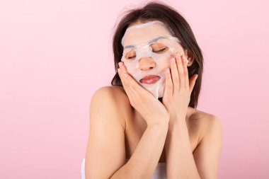 Çekici bir genç kız sadece beyaz bir havluya sarılıp yüz maskesi takıyor. Pembe arka planda izole edilmiş. Yakın plan..