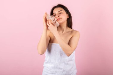 Çekici bir genç kız sadece beyaz bir havluya sarılmış bir yüz maskesi çıkarıyor. Pembe bir arkaplanda izole.