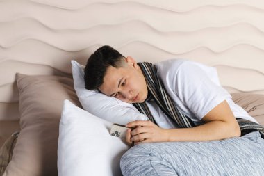 Genç adam bir eşarba sarılı olarak yatakta yatıyor ve hastalıktan yüzü kıpkırmızı olmuş, soğuk algınlığından hasta. Kötü hissediyorum, grip, ateş. telefon kullanarak ya da tutarak.