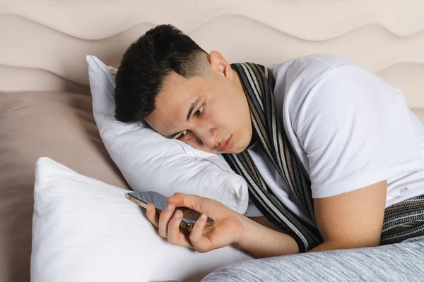 Genç adam bir eşarba sarılı olarak yatakta yatıyor ve hastalıktan yüzü kıpkırmızı olmuş, soğuk algınlığından hasta. Kötü hissediyorum, grip, ateş. telefon kullanarak ya da tutarak.