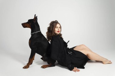 Boynunda zinciri olan genç, güzel bir doberman ve sahibi, uzun siyah ceketli, parlak kırmızı rujlu zarif bir kız. Beyaz bir arkaplanda izole edilmiş.