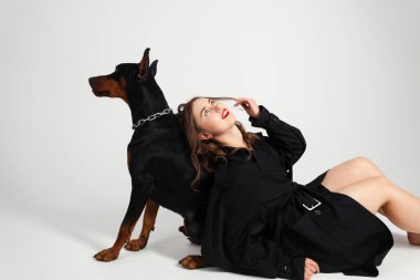 Boynunda zinciri olan genç, güzel bir doberman ve sahibi, uzun siyah ceketli, parlak kırmızı rujlu zarif bir kız. Beyaz bir arkaplanda izole edilmiş.