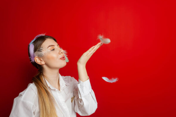 Portrait of a lovely tender girl wearing a feather decorations and blows away feathers from her hands. Isolated on a red background.