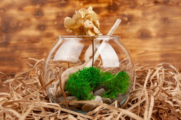 A glass bowl or vase filled with smooth rocks, flower and moss or plants standing on a hay against a wooden background.