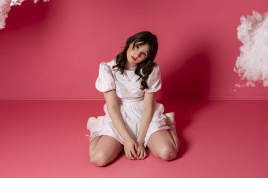 Süslü, beyaz bir elbise ve parlak bir makyaj giymiş sevimli, şık esmer kız bulutlara karşı bir bebek ya da kukla gibi poz veriyor ya da taklit ediyor. Pembe arka planda izole edilmiş.