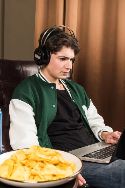 A boy or young man sitting on a couch and playing video games with a laptop while wearing headphones.  A table next to him with chips, soda, and energy drinks. Gamer.
