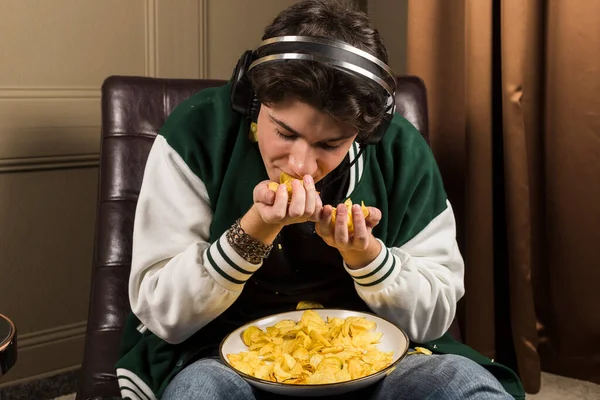 A gamer sitting on a couch and playing video games with a potato chips scattered all around him. An unkempt young man or boy. Dirty hands covered in oil.