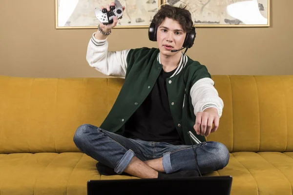 A boy or young man sitting on a couch or sofa and playing video games with a controller while wearing headphones. anger after losing. A gamer is throwing a controller.