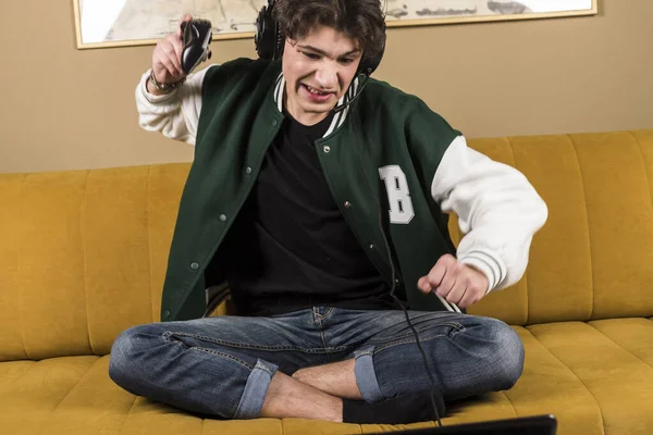 A boy or young man sitting on a couch or sofa and playing video games with a controller while wearing headphones. anger after losing. A gamer is throwing a controller.