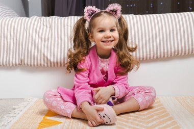 Pembe pijama ve bornozlu sevimli küçük bir kız yatağın önünde oturuyor ve mutlu bir şekilde gülümsüyor..