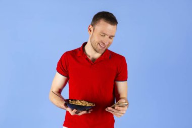 Kırmızı polo tişörtlü genç, yakışıklı, sağlıklı bir adam elinde bir kase mısır gevreği ya da kahvaltı tutuyor ve telefonuna bakıyor. Mavi arkaplanda izole.