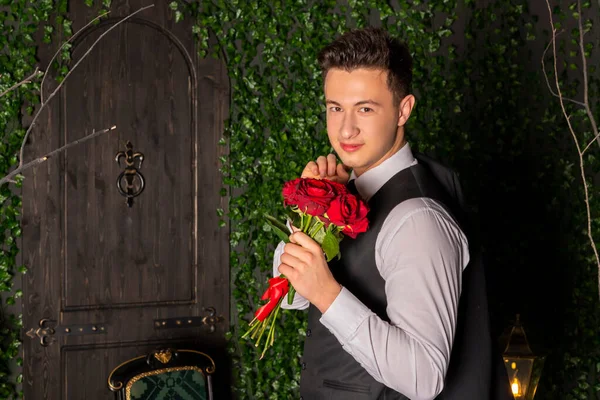 Portrait of an elegant young man in a fitted black formal suit holds red roses against an old vintage wall overgrown with vines. Formal or upscale event. Concept of elegance and sophistication
