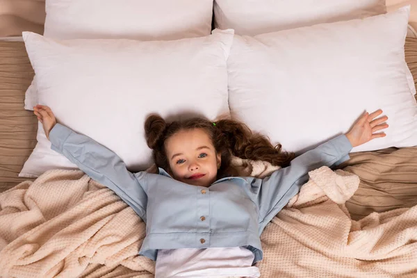 파란색 셔츠와 바지를 차려입은 매력적 소녀의 머리를 행복하게 베이지 침대에 — 스톡 사진