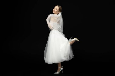 Güzel, alımlı bir kızın portresi, lüks ve zarif beyaz bir gelinlik içindeki gelin, elinde peçeyle nazikçe poz veriyor ve el kol hareketi yapıyor. Siyah bir arkaplanda izole edilmiş.