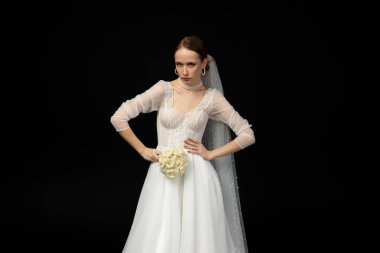 Güzel, çekici bir kız, lüks ve zarif beyaz bir gelinlik içinde zarif bir buket beyaz gülle, dikkatle, kötü niyetli ve ciddi görünüyor. Siyah bir arkaplanda izole edilmiş.
