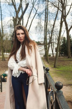 Parktaki bir derenin üzerindeki köprüde dikilen şık bir kız, omuzlarının üzerine atılmış bir palto, bileğinde akıllı bir saat, şık, koyu mavi pantolon, beyaz gömlek..