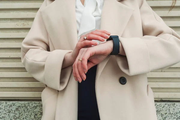 Fashionable unrecognizable girl in classic clothes standing over a facade of a building. light beige coat, looking at her smart watch, checks the time.