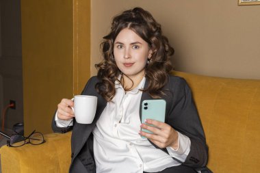 Güzel bir iş kadını, resmi olarak giyinmiş, elinde telefon ve fincanla şehirde oturuyor..