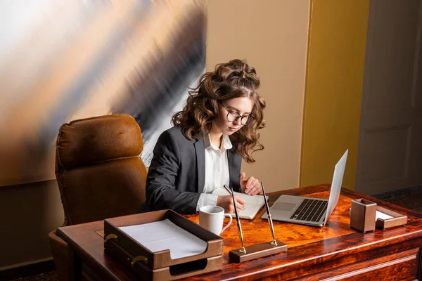 Stock image A beautiful businesswoman, formally dressed, sits in her office and working with a laptop and notebook. Concept of professionalism and productivity.