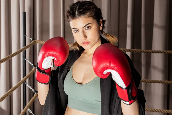 A beautiful confident girl in red boxing gloves stands in a fighting stance and looks into the camera in a boxing ring.