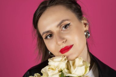 Deri ceket ve kot pantolon giyen havalı motorcu kız bir buket beyaz gülle poz veriyor. Pembe arka planda izole edilmiş.