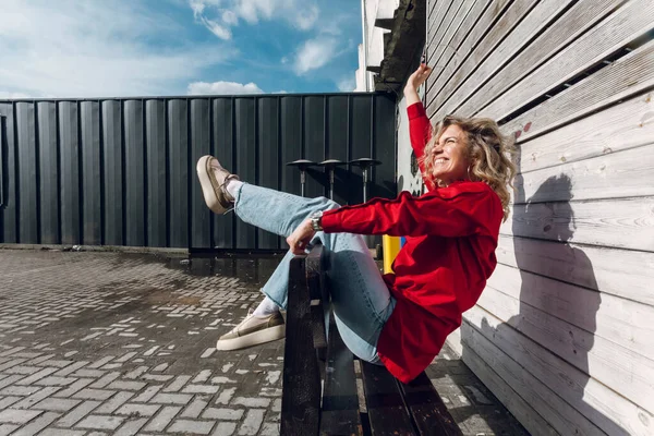 Street photography. A happy stylish curly blonde girl in red shirt posing on a street bench.