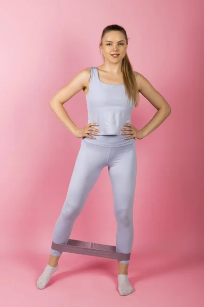 身穿灰色运动服的年轻健康女孩正在做伸展运动和带着橡皮筋抵制的热身运动 以粉红为背景隔离 — 图库照片