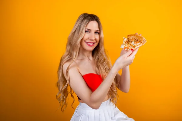 A beautiful sensual fit young blonde girl in a short top and skirt is sitting on a bar stool posing with a pizza slice. Isolated on yellow.