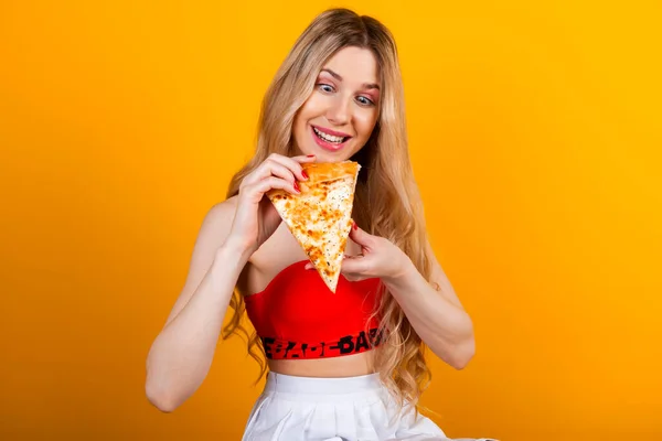 A beautiful sensual fit young blonde girl in a short top and skirt posing with a pizza slice. Isolated on yellow.