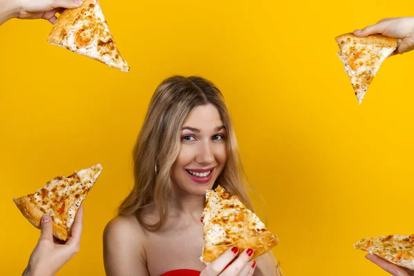 A beautiful sensual fit young blonde girl surrounded with hands with pizza slices around her. Isolated on yellow.